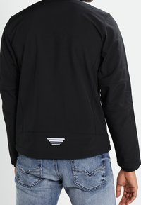 Black softshell jacket with a smooth texture, featuring a subtle logo at the back and tailored fit, paired with light blue jeans.