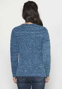 Un pull en tricot bleu avec une finition texturée, présentant des manches longues, un col montant et un ourlet légèrement évasé, porté sur un jean sombre.