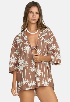 Camicia marrone con bottoni, caratterizzata da un motivo di palme bianche, maniche corte e una vestibilità rilassata. Il tessuto sembra leggero, con una texture simile al lino.