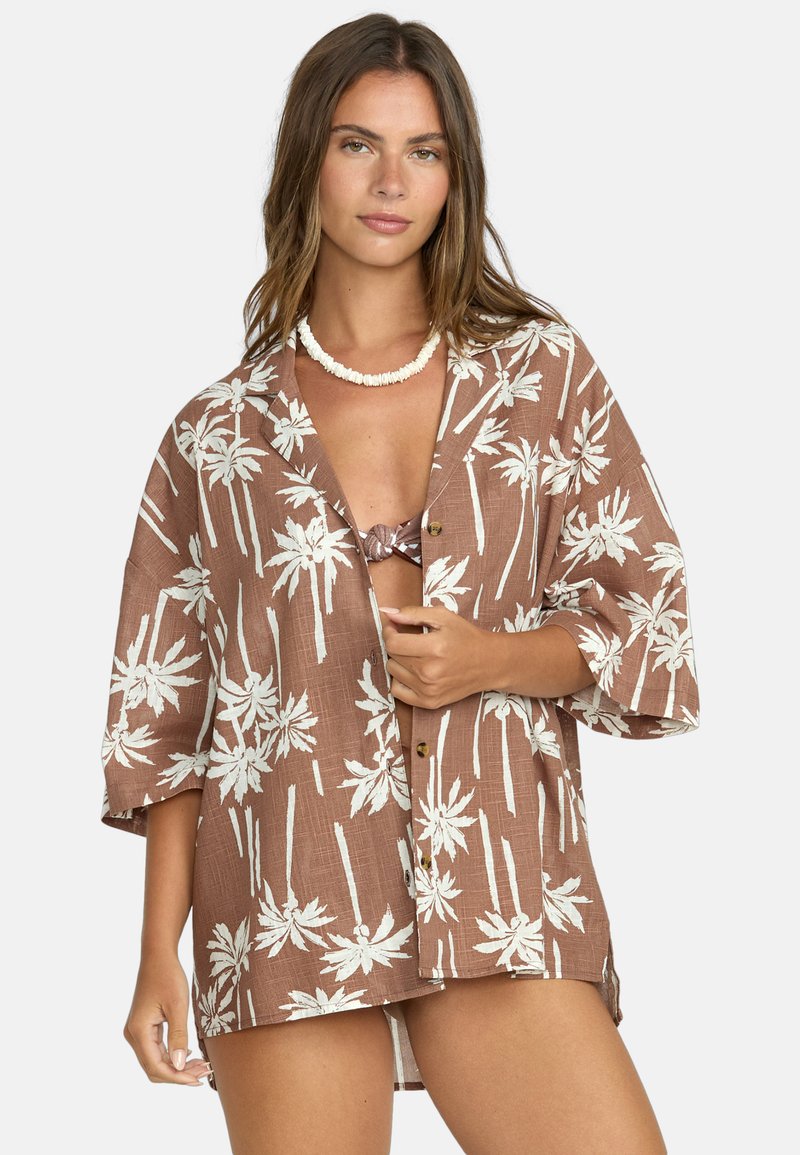 Camicia marrone con bottoni, caratterizzata da un motivo di palme bianche, maniche corte e una vestibilità rilassata. Il tessuto sembra leggero, con una texture simile al lino.