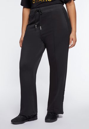 Osoba ubrana w czarne spodnie ze sznurkiem i czarne buty sportowe, stojąca na tle jednolitego białego tła.