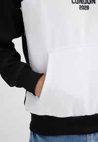 Sudadera con capucha de manga negra y cuerpo blanco, que cuenta con un bolsillo canguro, puños de canalé y "LONDRES 2028" en letras negritas.
