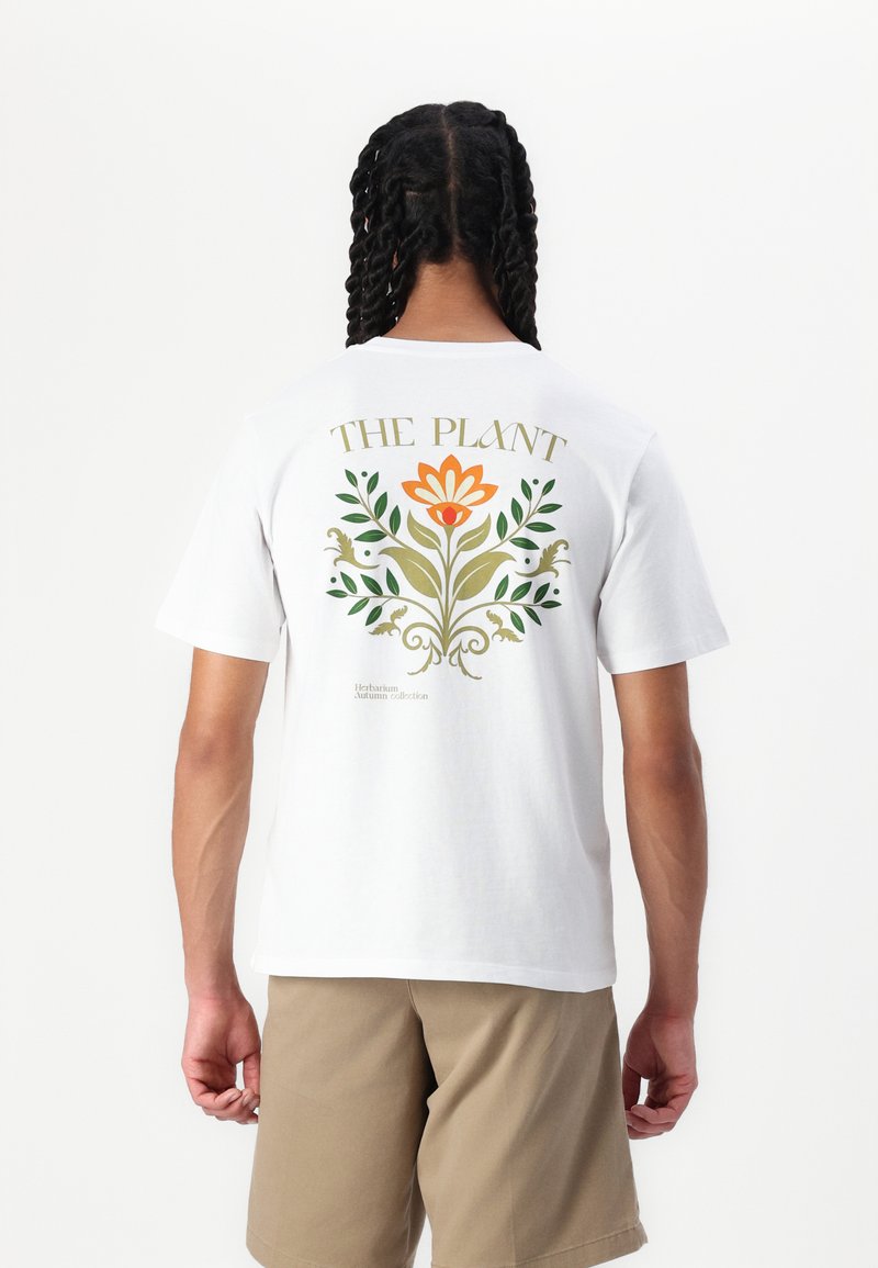 Tricou alb din bumbac cu un design floral în portocaliu și verde pe spate, având textul "PLANTA" deasupra și accente botanice dedesubt.