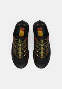 Μαύρα αθλητικά παπούτσια με κίτρινες και κόκκινες λωρίδες στα κορδόνια, με υφή στο επάνω μέρος και μαλακές σόλες, σχεδιασμένα για απόδοση και άνεση.