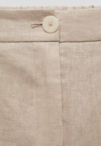 Béžová lněná tkanina s knoflíkovým zapínáním a sklady kolem pasu. Textura je mírně hrubá, což vytváří přirozený a uvolněný vzhled.