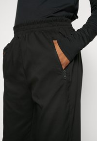 Black athletic pants featuring an elastic waistband, zippered side pockets, and a smooth, slightly textured fabric. Detail stitching visible.