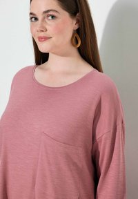 Hellrosa Langarmshirt mit weiter Ausschnittform und einer Brusttasche. Weicher Stoff mit entspannter Passform und abgesenkten Schulternähten.