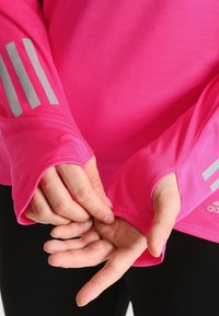 Ręce regulujące otwory na kciuki w jasnoróżowej, długiej koszulce sportowej z odblaskowymi paskami i logo Adidas na rękawach.