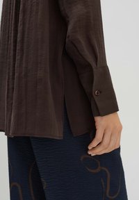 Brown, textured shirt with side slits and a buttoned cuff, paired with a dark blue, patterned garment featuring wavy lines.