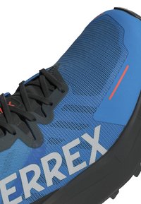 Blå löparskor med en texturerad meshöverkonstruktion, svarta accenter och grå varumärkning. Anmärkningsvärda detaljer inkluderar tunna svarta snören och klara orangea höjdpunkter.