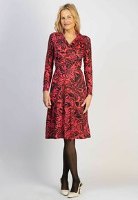 Rotes Kleid mit langen Ärmeln, versehen mit einem schwarzen Blumenmuster, V-Ausschnitt und taillierter Passform, die in einen Rock übergeht. Getragen mit schwarzen Strumpfhosen und High Heels.