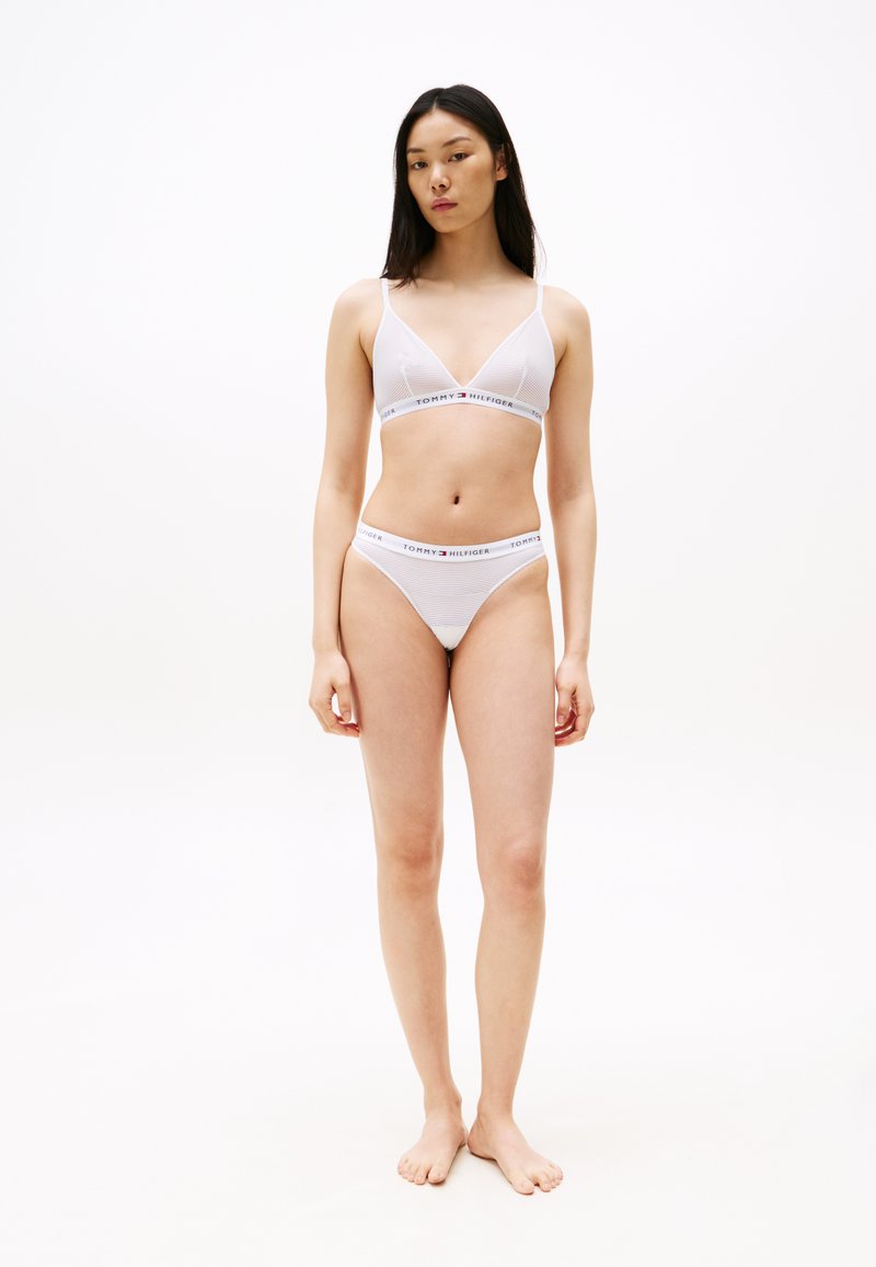 Hvit undertøysett som består av en bralette med trekantede kopper og matchende stringtruse. Begge har et logobånd med alternerende striper. Glatt tekstur.
