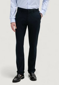 Pantaloni tailored blu navy scuri con una linea elegante, caratterizzati da una texture liscia e una chiusura con bottone, abbinati a mocassini in pelle nera.