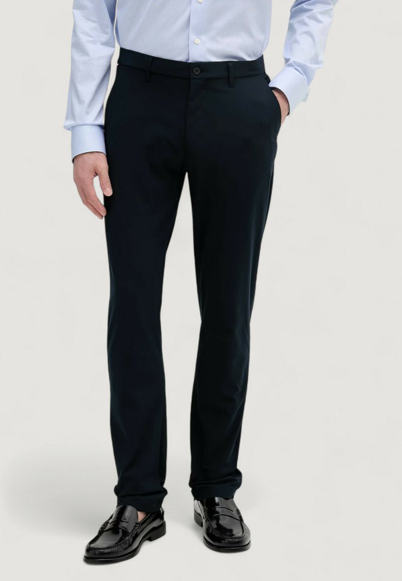 Pantaloni tailored blu navy scuri con una linea elegante, caratterizzati da una texture liscia e una chiusura con bottone, abbinati a mocassini in pelle nera.