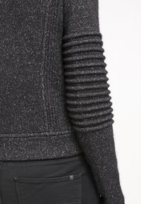 Primer plano de un suéter de punto gris oscuro con detalles acanalados y acolchados en la manga, combinado con pantalones negros que muestran un bolsillo trasero.