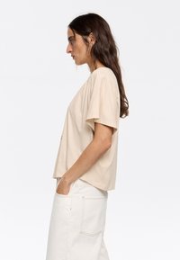 T-shirt beige a maniche corte realizzato in tessuto morbido, con un taglio morbido e scollo rotondo, abbinato a pantaloni bianchi a vita alta.
