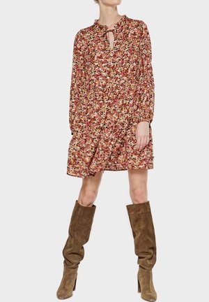 Blumenkleid mit einem Schlüssel-Ausschnitt, langen Ärmeln und geraffter Taille. Hat ein Muster in Rosa, Creme und Grün. Kombiniert mit kniehohen, braunen Stiefeln.