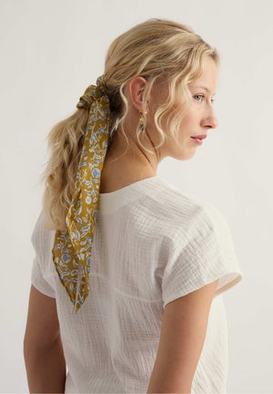 Szőke nő laza fürtökkel, lófarokba kötve egy mustárszínű, virágos sállal, fehér, texturált rövid ujjú felsőt viselve, jobbra nézve.