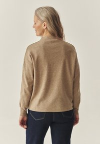 Béžový pletený sveter s vysokým golierom, s detailom zadných gombíkov. Kombinovaný s tmavými rifľovými džínsami. Mäkká textúra, pohodlný strih.