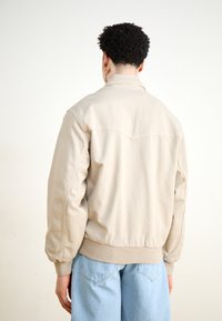 Beige bomber dzseki laza szabással, bordás mandzsettával és aljjal. Lekerekített gallérral és sima textúrával rendelkezik, amelyhez világoskék farmernadrágot visel.