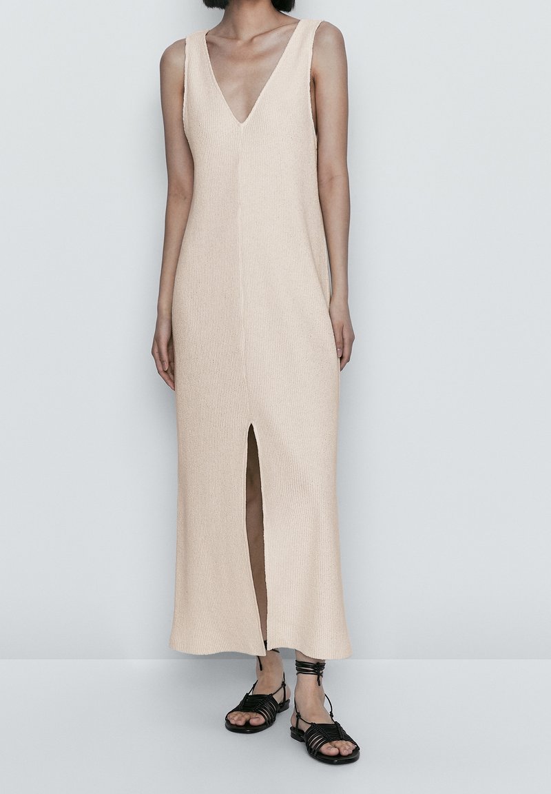 Femme portant une robe midi beige en maille sans manches avec une fente à l'avant et des sandales plates noires à lanières, debout devant un fond uni.