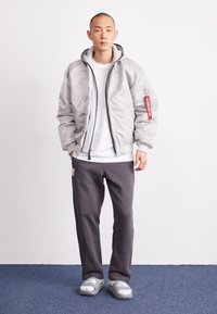 Hellgraue Bomberjacke mit einem Frontreißverschluss, gerippten Bündchen und einer angenähten Kapuze, kombiniert mit dunkelgrauen Jogginghosen und grauen Rutsch-Sandalen.