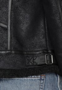 Czarna skórzana kurtka z teksturowanym wykończeniem, z klamrą paska i kontrastowym futrem owczym przy brzegach.