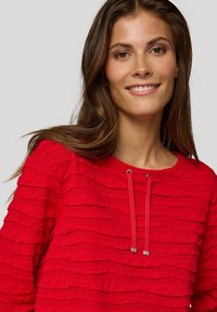 Junge Frau mit langen braunen Haaren, die lächelt und einen strukturierten, leuchtend roten Pullover mit Kordelzug trägt, vor einem schlichten, hellen Hintergrund.