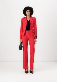 Blazer et pantalon sur mesure rouges, associés à un top noir. Des escarpins noirs à bouts pointus et un petitSac à main noir complètent le look. Texture lisse.