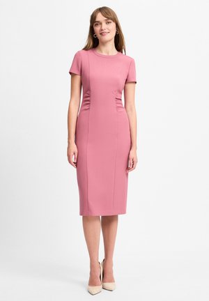 Vrouw die een nauwsluitende stofroze jurk tot op de knieën draagt met korte mouwen en beige pumps, staand tegen een witte achtergrond.