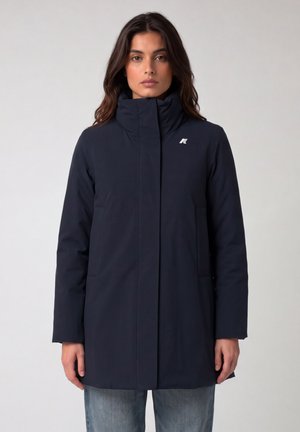 Donna con giacca blu scuro a collo alto, maniche lunghe e tasche frontali, abbinata a jeans azzurri su sfondo semplice.