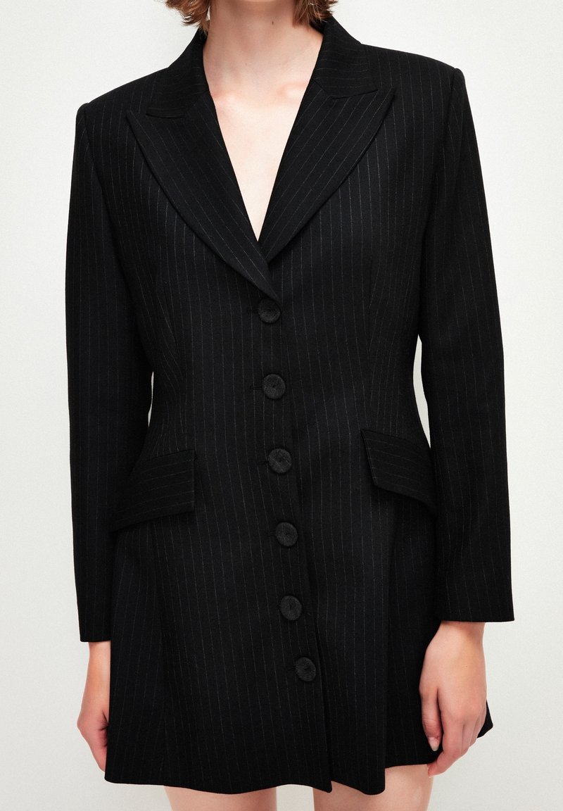 Femme portant une robe blazer à fines rayures noires, manches longues, col cranté, boutons sur le devant et poches à rabat.