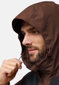 Giacca cappuccio marrone in tessuto liscio e resistente all'acqua. Caratterizzata da chiusura con cordino e accenti scuri a contrasto sul colletto.