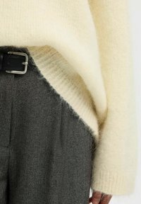 Kreminės spalvos pūkuota megztinis ant pilkų tekstūruotų kelnių. Megztinis turi įrišimo apdailą, o kelnės prisispaustos juodu odiniu diržu.