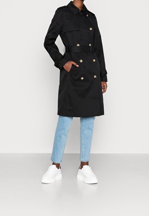 Personne portant un trench-coat noir à double boutonnage avec ceinture, un jean bleu clair et des baskets blanches, debout devant un fond uni.