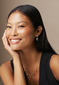 Gold jewelry including a chain necklace with charms, a star-shaped stud earring, and a gemstone drop earring. Smooth skin and dark hair.
