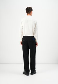 Weißes langärmeliges Hemd mit strukturiertem Muster, kombiniert mit schwarzen Hosen. Verfügt über sichtbare Taschen und ein blaues Etikett am Bund. Schwarze Schuhe.