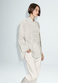 Camicia a righe grigio chiaro con colletto alto e maniche lunghe, caratterizzata da una vestibilità rilassata e un orlo asimmetrico, abbinata a pantaloni beige chiaro.