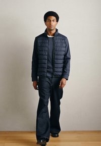 Marineblå puffervest over en ribbet mørk sweater, kombineret med vidde-ben denimjeans og sorte sko, mod en neutral baggrund.