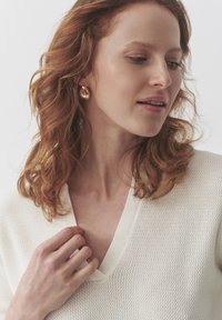 Haut en tricot crème avec un décolleté en V et un motif texturé. Un modèle a des cheveux roux ondulés jusqu'aux épaules et porte des boucles d'oreilles en or.