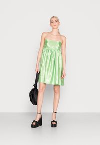 Vestido de cetim verde com corpete justo, cintura franzida e saia godê. A modelo segura uma bolsa preta e usa sapatos plataforma pretos e robustos.