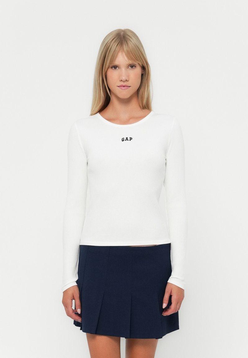 Witte ribbel trui met lange mouwen en een ronde halslijn, met een zwart "GAP" logo in het midden, gecombineerd met een marineblauwe geplooide rok.