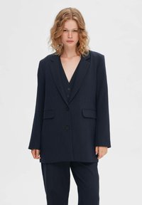 Marineblauwe op maat gemaakte blazer met lange mouwen, notch revers en twee voorzakken, gemaakt van een licht gestructureerde stof en voorzien van een enkele knoopsluiting.
