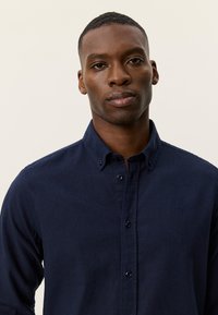 Πουκάμισο σε ναυτικό μπλε χρώμα με κουμπιά, κατασκευασμένο από υφασμάτινη υφή, με μικρό λογότυπο, κανονικό γιακά και μακριά μανίκια με μανσέτες κουμπιών.