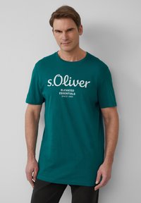 Turkusowa koszulka z krótkim rękawem o okrągłym dekolcie, z białym napisem: "s.Oliver Elevated Essentials Since 1969." Miękka tkanina, luźny krój.