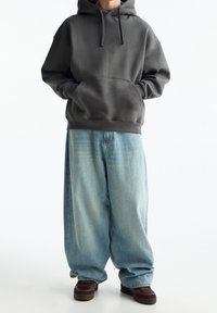Sudadera con capucha de color gris carbón con un bolsillo tipo canguro, combinada con unos vaqueros de denim sueltos y de color azul claro, y zapatos de color marrón oscuro.