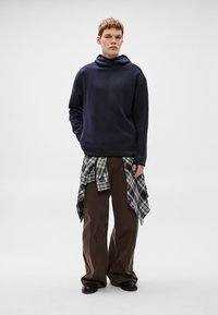 Námořnická mikina s vysokým límcem, volné hnědé kalhoty a kostkovaná košile uvázaná kolem pasu. Hladká textura, neformální design.