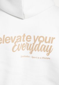 Hoodie blanche avec du texte brun qui lit "Élevez votre quotidien" et "Smilodox - Le sport est un style de vie." Texture de tissu douce, design décontracté.