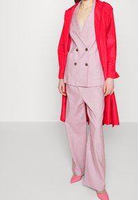 Czerwony trencz na różowym dwurzędowym żakiecie i dopasowanych szerokich spodniach. Różowe czółenka na wysokim obcasie dopełniają stylizację. Gładkie tekstury w całym wyglądzie.