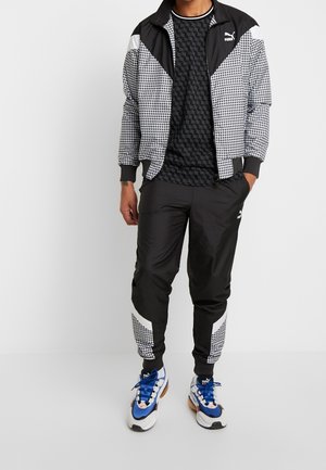 Homme portant une veste de survêtement Puma noire et blanche avec un motif géométrique, un pantalon assorti, une chemise noire à motif et des baskets blanches avec des lacets bleus.